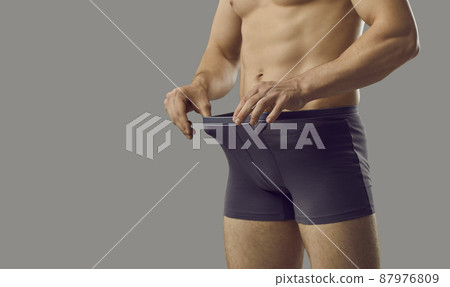 Handsome young male looks inside underpants at his private area. Shirtless attractive muscular man in underwear briefs looking at his penis standing isolated on grey background. Genital health concept Handsome young male looks inside underpants at his private area. Shirtless attractive muscular man in underwear briefs looking at his penis standing isolated on grey background. Genital health concept 87976809