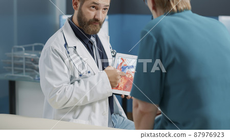 General practitioner holding tablet with heart condition and cardiology illustration at consultation with old patient. Doctor explaining cardiovascular system and diagnosis to elder woman. 87976923