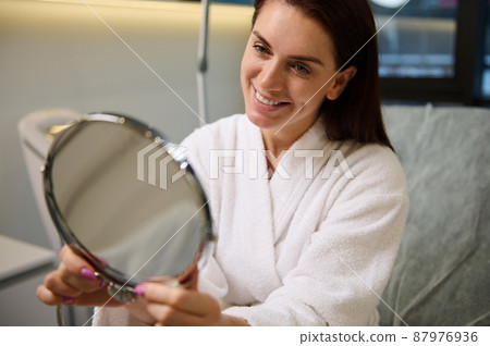 Attractive middle aged European woman in white bathrobe smiles while looking at her mirror reflection, admiring her appearance. Beauty, skin and body care, hygiene, cosmetology and spa concept Attractive middle aged European woman in white bathrobe smiles while looking at her mirror reflection, admiring her appearance. Beauty, skin and body care, hygiene, cosmetology and spa concept 87976936