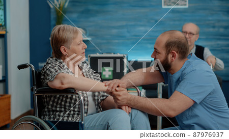 Man nurse holding hands and comforting pensioner with disability in nursing home. Medical assistant having conversation with elder disabled woman about rehabilitation and healthcare. 87976937