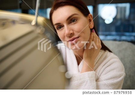 Beautiful middle aged woman in white terry bathrobe looking at her reflection in a round cosmetic mirror, admiring her appearance. Beauty, skin and body care, hygiene, cosmetology and spa concept 87976938