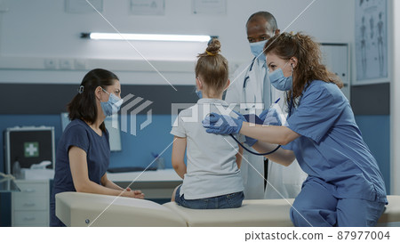 Woman nurse using stethoscope to check pulse and heartbeat at checkup visit. Medical assistant with face mask consulting sick little girl at examination appointment in cabinet. Woman nurse using stethoscope to check pulse and heartbeat at checkup visit. Medical assistant with face mask consulting sick little girl at examination appointment in cabinet. 87977004