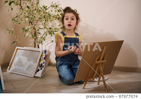 Charming little girl in yellow t-shirt and blue denim holding a paintbrush and painting on canvas using watercolor or oil paints, sitting on floor next to painted pictures at home. Kids art concept 87977074