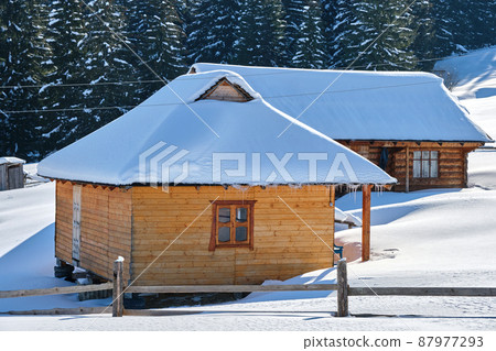 Small wooden house covered with fresh fallen snow surrounded with tall pine trees in winter mountains. Small wooden house covered with fresh fallen snow surrounded with tall pine trees in winter mountains. 87977293