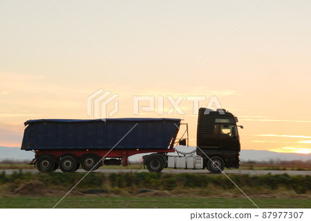 Semi-truck with tipping cargo trailer transporting sand from quarry driving on highway hauling goods in evening. Delivery transportation and logistics concept Semi-truck with tipping cargo trailer transporting sand from quarry driving on highway hauling goods in evening. Delivery transportation and logistics concept 87977307