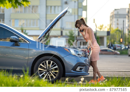Helpless woman standing near her car with open hood takling on mobile phone calling road service for help. Young female driver having trouble with vehicle. Helpless woman standing near her car with open hood takling on mobile phone calling road service for help. Young female driver having trouble with vehicle. 87977585