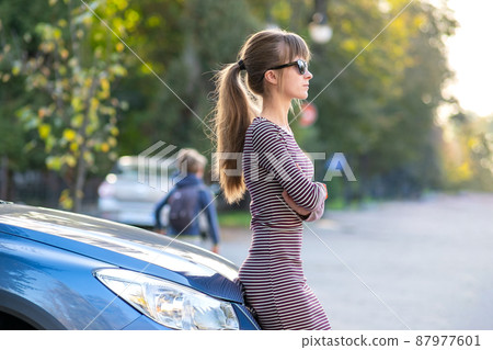 Happy young woman driver enjoying warm summer day standing beside her car on a city street. Travelling and vacation concept. Happy young woman driver enjoying warm summer day standing beside her car on a city street. Travelling and vacation concept. 87977601