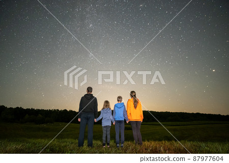 Happy family standing in night field looking at dark sky with many bright stars. Parents and children observing meteor shower. 87977604