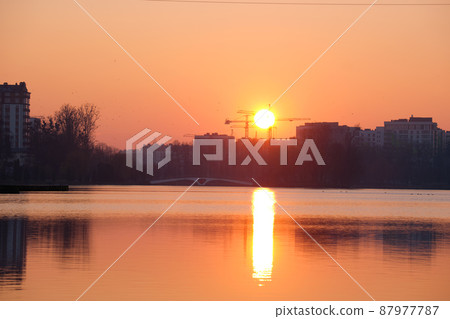 Dark silhouette of tower cranes with big setting sun at high residential apartment buildings construction site at sunset on lake shore. Real estate development 87977787