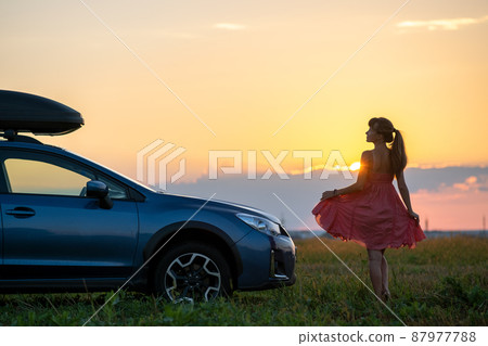 Dark silhouette of lonely woman relaxing near her car on grassy meadow enjoying view of colorful sunrise. Young female driver resting during road trip beside SUV vehicle. 87977788