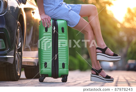 Closeup of young female legs sitting on suitcase bag beside car. Travelling and vacations concept. Closeup of young female legs sitting on suitcase bag beside car. Travelling and vacations concept. 87978243