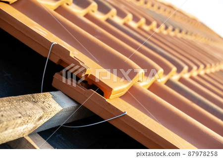 Closeup of metal montage anchor for installation of yellow ceramic roofing tiles mounted on wooden boards covering residential building roof under construction. 87978258