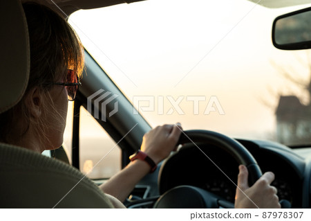 Close up view of a woman driver holding steering wheel driving a car at sunset. Close up view of a woman driver holding steering wheel driving a car at sunset. 87978307