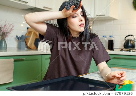 Tired young woman with a metal sponge for cleaning, washes a baking sheet in the kitchen 87979126