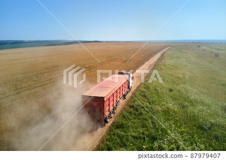 Aerial view of cargo truck driving on dirt road between agricultural wheat fields making lot of dust. Transportation of grain after being harvested by combine harvester during harvesting season Aerial view of cargo truck driving on dirt road between agricultural wheat fields making lot of dust. Transportation of grain after being harvested by combine harvester during harvesting season 87979407