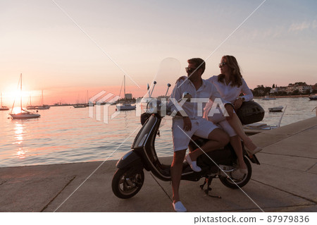The couple enjoys a vacation while sitting on a scooter by the sea and enjoying the beautiful sunrise. Selective focus  87979836