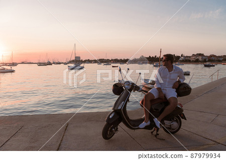 A modern senior businessman in casual clothes sitting on a scooter by the sea and enjoys the sunrise. Selective focus A modern senior businessman in casual clothes sitting on a scooter by the sea and enjoys the sunrise. Selective focus 87979934