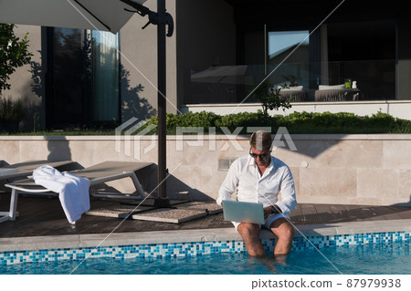 A modern elderly man enjoys the pool while working on his laptop next to a modern luxury house. Selective focus  87979938