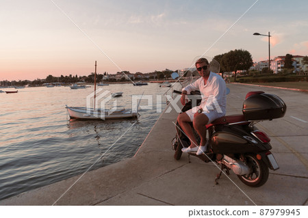 A modern senior businessman in casual clothes sitting on a scooter by the sea and enjoys the sunrise. Selective focus A modern senior businessman in casual clothes sitting on a scooter by the sea and enjoys the sunrise. Selective focus 87979945