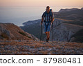 Young hiker with backpack and trekking poles walking along a mountain path at sunset with a smile. High quality photo. 87980481