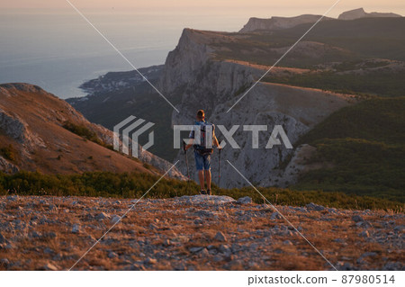 Young hiker with backpack and trekking poles enjoying the scenery at sunset with a smile. High quality photo. 87980514