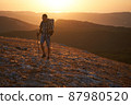 Young hiker with backpack and trekking poles walking along a mountain path at sunset with a smile. High quality photo. 87980520