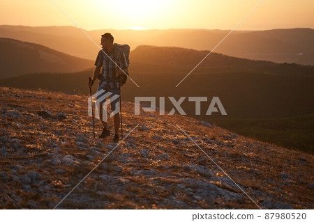 Young hiker with backpack and trekking poles walking along a mountain path at sunset with a smile. High quality photo. 87980520