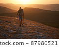 Young hiker with backpack and trekking poles walking along a mountain path at sunset with a smile. High quality photo. 87980521