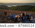 Young hiker with backpack and trekking poles taking rest and enjoying the scenery at sunset with a smile. High quality photo. 87980524