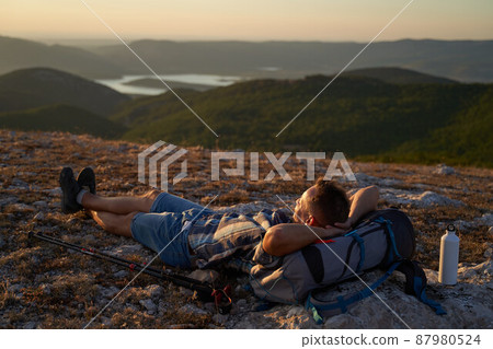 Young hiker with backpack and trekking poles taking rest and enjoying the scenery at sunset with a smile. High quality photo. 87980524
