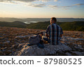 Young hiker with backpack and trekking poles taking rest and enjoying the scenery at sunset with a smile. High quality photo. 87980525