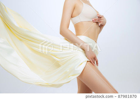 Portrait of slim female body in beige cotton lingerie covered with silk flying fabrik isolated over gray studio background 87981870
