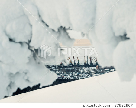 Winter northern seaport. View through the snowy branches. Cargo seaport in the Arctic. Large harbor cranes on distance a background of ice and snow. Industry and transport infrastructure in the North 87982907