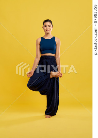 Front view of flexible girl practicing yoga pose at studio. Brunette female standing on one leg, holding foot by hand, with mudras, looking at camera, smiling. Concept of yoga. Front view of flexible girl practicing yoga pose at studio. Brunette female standing on one leg, holding foot by hand, with mudras, looking at camera, smiling. Concept of yoga. 87983359