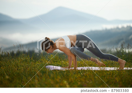 Side panoramic view of young woman practicing yoga in hills. Sporty girl standing on hands with knee raised on yoga mat in nature. Concept of healthy living. 87983380