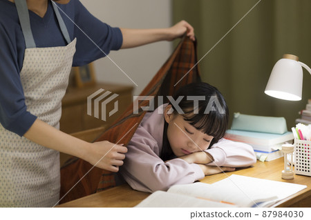 A mother who puts a blanket on her daughter who falls asleep at the desk 87984030