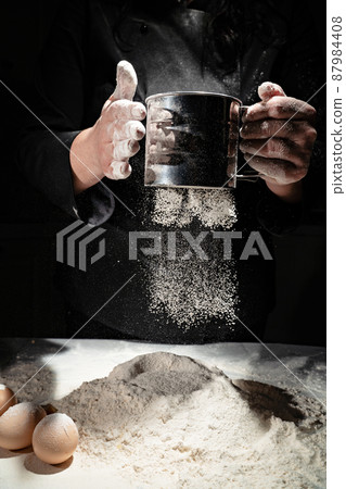 woman sifts flour through sieve onto table with eggs on black background.  87984408
