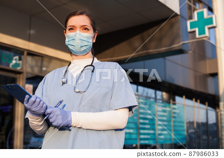 Young female doctor standing in front of healthcare facility, wearing protective face mask and PPE equipment, holding medical patient clipboard, COVID-19 pandemic crisis Young female doctor standing in front of healthcare facility, wearing protective face mask and PPE equipment, holding medical patient clipboard, COVID-19 pandemic crisis 87986033