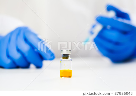 Yellow ampoule vaccine liquid vial on table and hands in blue protective medical gloves holding syringe jab 87986043