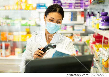 Portrait of female pharmacist in medical mask working at the cash register in pharmacy - scans barcode on medicine package 87992722