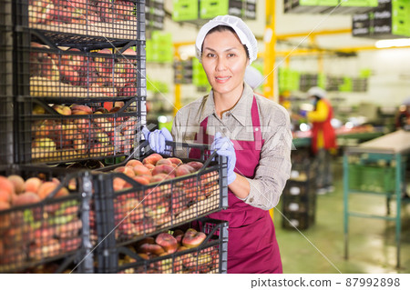 Glad woman in uniform holding crate with peaches during packaging 87992898