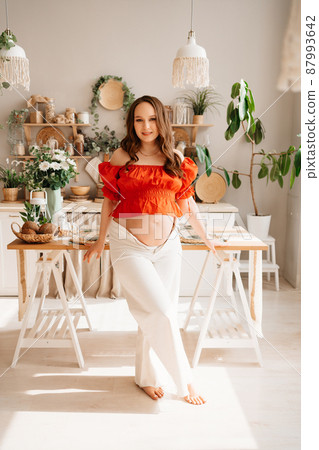 a beautiful pregnant woman in a romantic orange blouse and trousers in kitchen.  87993642