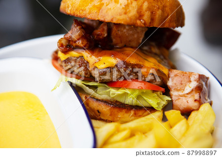 The concept of fast food and takeaway food. A juicy hamburger lies next to french fries on a metal plate along with cheddar cheese sauce in the background. 87998897