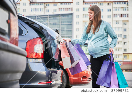 Side view of Caucasian woman with belly puting shopping bags in automobile trunk enjoying maternity and parenthood in awaiting mode. Pregnant female preparing for baby birth. 87999781