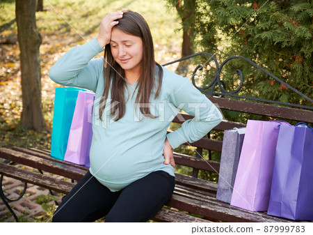 Surprised pregnant woman sitting on street bench, putting one hand on her forehead and other on waist. Expectant mother resting on wooden bench after shopping, putting packages next to each other. 87999783