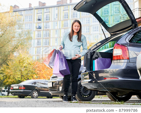 Pregnant woman with paper bags give premature child birth at auto parking in city downtown. Young woman dressed in casual wear feeling pain during broken water after shopping for awaiting baby 87999786