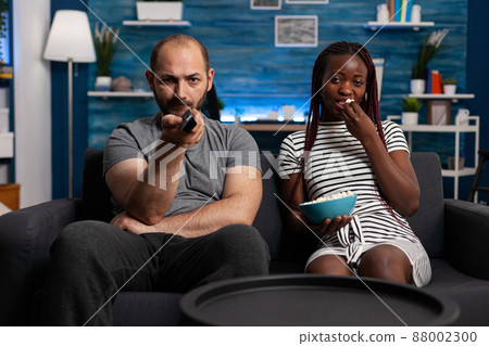 Caucasian man and african american woman watching TV while sharing a bowl of popcorn sitting on sofa at home. Stressed husband angrily switching television programs looking for a movie. Caucasian man and african american woman watching TV while sharing a bowl of popcorn sitting on sofa at home. Stressed husband angrily switching television programs looking for a movie. 88002300