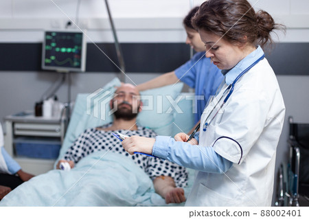 Doctor with stethoscope reading medical chart while nurse is making patient comfortable after surgical intervention. Middle aged man recovering lying in hospital bed attended by team of doctors. Doctor with stethoscope reading medical chart while nurse is making patient comfortable after surgical intervention. Middle aged man recovering lying in hospital bed attended by team of doctors. 88002401