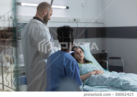 Medical doctor and nurse doing consult in private ward to recovering woman in hospital bed after surgical intervention. Patient with nasal cannula looking at health care professionals. 88002425