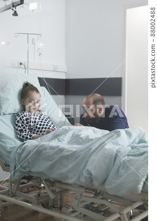 Anxious sad man watching unwell little daughter sleeping in clinic bed while in pediatric hospital patient ward. Under treatment sedated ill child resting while attentive parent taking care of her. Anxious sad man watching unwell little daughter sleeping in clinic bed while in pediatric hospital patient ward. Under treatment sedated ill child resting while attentive parent taking care of her. 88002488